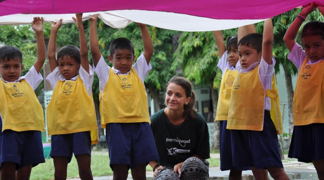 Projects Abroad volunteer under a parachute with children during an outdoor activity at a kindergarten in Thailand. A volunteer working with children in Thailand crouches under a parachute during an activity with kids at her Childcare placement.